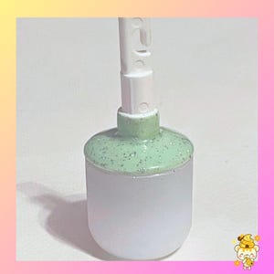 May include: A close-up of a white and green toothbrush head. The toothbrush head has a cylindrical shape with a light green, speckled top. The handle is white and has a few small holes.