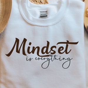 Può includere: Felpa bianca con il testo "Mindset is everything" in lettere corsive marroni.