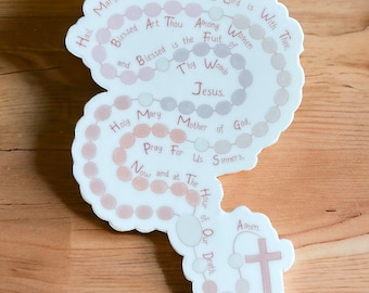 May include: A white sticker with a pink and gray rosary design. The sticker features the traditional Catholic prayer, "Hail Mary, full of Grace..." and "Holy Mary, Mother of God..." The sticker also includes a cross at the bottom.