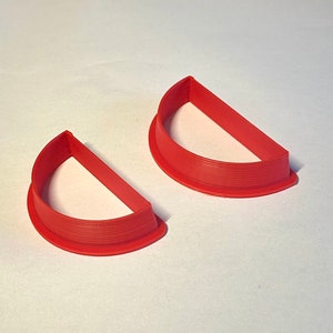 Puede incluir: Dos cortadores de galletas de plástico rojo en forma de semicírculos. Los cortadores tienen un borde elevado alrededor del perímetro.