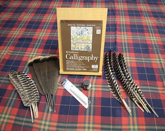 May include: A calligraphy set featuring a pad of Strathmore Calligraphy writing paper, several feathers, a bottle of black ink, and a pen nib. The items are arranged on a red, blue, and yellow plaid fabric.