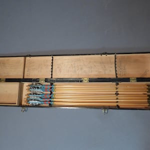 May include: A wooden box with a black exterior and a hinged lid. The box is open and contains a set of 12 arrows with blue, red, and yellow fletching. The arrows are arranged in a row and are all the same length.