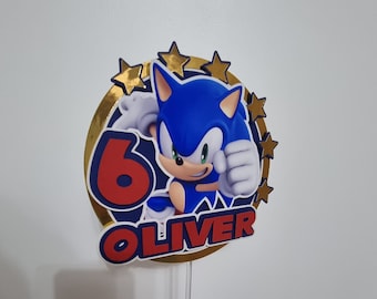 May include: A blue and red Sonic the Hedgehog cake topper with a gold border and five gold stars. The topper says "6 Oliver" in red letters.