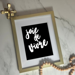 Puede incluir: Una impresi&oacute;n art&iacute;stica enmarcada con la frase "joie de vivre" en escritura blanca sobre un fondo negro. El marco es dorado y la impresi&oacute;n est&aacute; enmarcada con un borde blanco. Cuentas de madera decorativas y un objeto geom&eacute;trico dorado tambi&eacute;n est&aacute;n en la imagen.