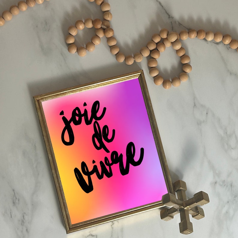 Puede incluir: Una impresi&oacute;n enmarcada con las palabras "joie de vivre" en escritura negra sobre un vibrante fondo degradado de amarillo, rosa y morado. El marco es dorado, y tambi&eacute;n hay una guirnalda decorativa de cuentas de madera y un objeto geom&eacute;trico dorado.
