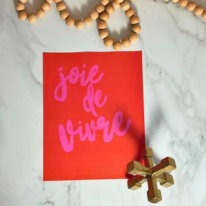Puede incluir: Un cuadrado rojo con las palabras "joie de vivre" en escritura rosa. Un jack de metal dorado y una hebra de cuentas de madera tambi&eacute;n est&aacute;n en la imagen. Los art&iacute;culos est&aacute;n dispuestos sobre una superficie de m&aacute;rmol blanco.