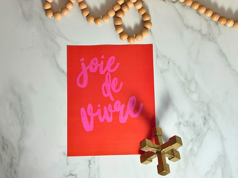 Puede incluir: Un cuadrado rojo con las palabras "joie de vivre" en escritura rosa. Un jack de metal dorado y una hebra de cuentas de madera tambi&eacute;n est&aacute;n en la imagen. Los art&iacute;culos est&aacute;n dispuestos sobre una superficie de m&aacute;rmol blanco.