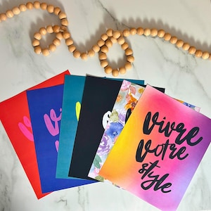 Puede incluir: Una colecci&oacute;n de tarjetas de felicitaci&oacute;n coloridas con varios dise&ntilde;os y texto. Las tarjetas son rojas, azules, turquesas y negras, con patrones florales y de acuarela. Una tarjeta presenta la frase "Vivre Votre Style" en escritura negra.