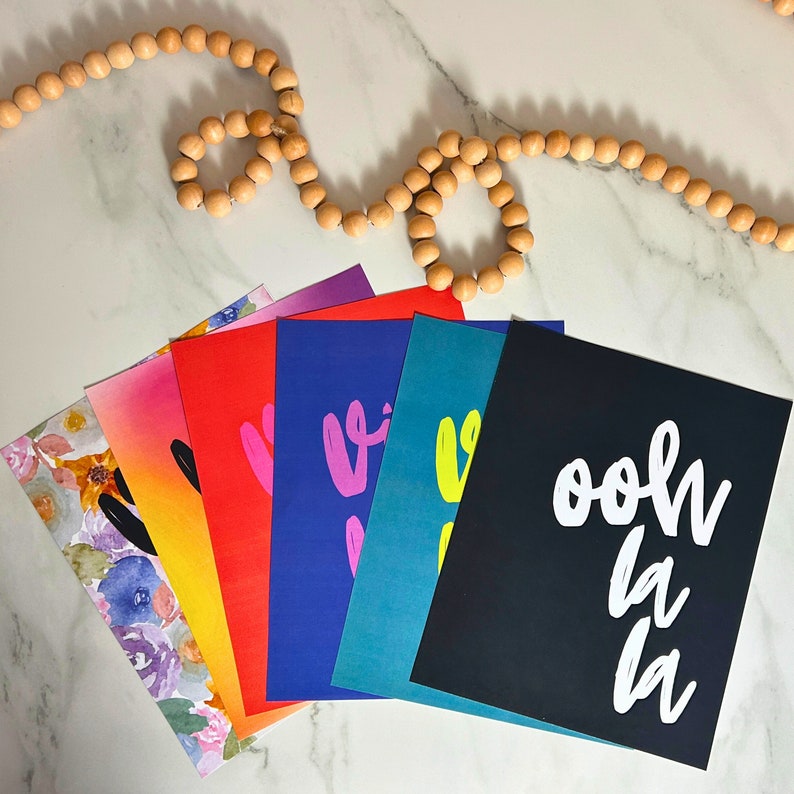 Puede incluir: Una colecci&oacute;n de tarjetas de felicitaci&oacute;n coloridas abanicadas sobre una superficie de m&aacute;rmol blanco. Las tarjetas presentan dise&ntilde;os florales en acuarela y texto audaz como "ooh la la" y "vibe" en varios colores, incluyendo rosa, amarillo y negro. Una guirnalda de cuentas de madera est&aacute; en el fondo.