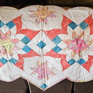 May include: A white quilted table runner with a geometric pattern of red, blue, and pink floral designs. The runner has a scalloped edge and is perfect for adding a touch of elegance to any table.