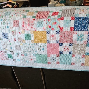 May include: A long, rectangular quilt with a patchwork design. The quilt is made up of small squares of fabric in various colours and patterns, including red, blue, green, yellow, and white. The quilt is edged with a light blue trim.