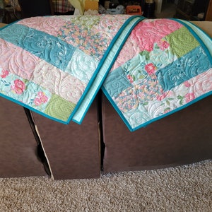 May include: Two quilted sofa arm covers with a teal border. The covers are made of patchwork fabric with floral patterns in shades of pink, green, and blue.