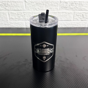 Puede incluir: Un vaso térmico negro con tapa transparente y pajita negra. El vaso tiene un emblema plateado con el texto "THE SHEARED RAM LUMBRIDGE PREMIUM LUMBRIDGE ALE". Está diseñado para mantener las bebidas frías o calientes.