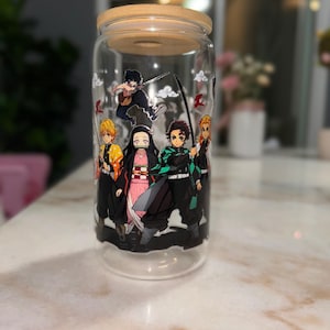 May include: A clear glass jar with a wooden lid, featuring a colorful anime-style graphic of several characters. The jar is cylindrical and appears to be empty. The characters are depicted with swords and various outfits.