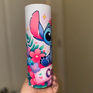 May include: A white tumbler featuring a colorful cartoon character with large ears, blue fur, and a floral crown. The tumbler is decorated with pink, blue, and purple flowers, and the name "Co" is visible on a banner.