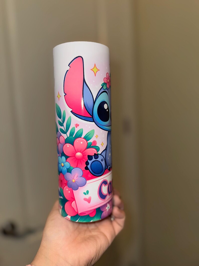 May include: A white tumbler featuring a colorful cartoon character with large ears, blue fur, and a floral crown. The tumbler is decorated with pink, blue, and purple flowers, and the name "Co" is visible on a banner.