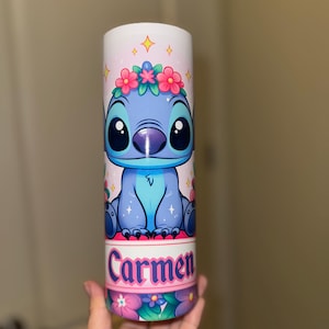 May include: A cylindrical tumbler featuring a cartoon character with a blue body, large black eyes, and a floral crown. The tumbler has the name "Carmen" printed in pink and white, with a floral pattern at the base.