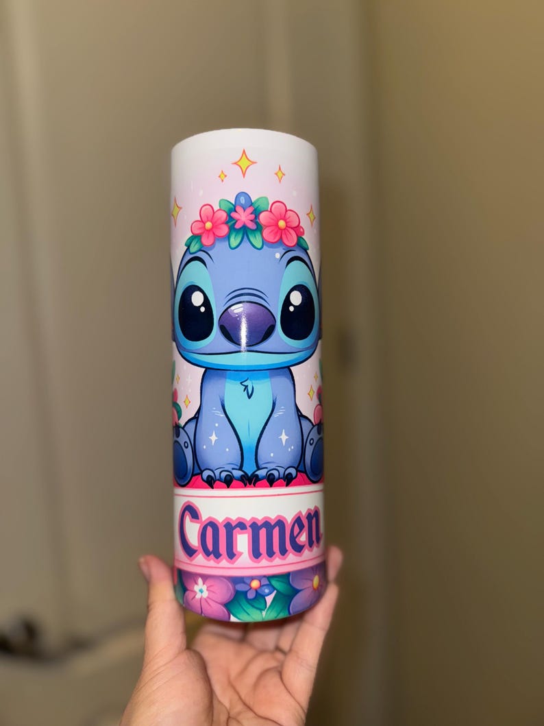 May include: A cylindrical tumbler featuring a cartoon character with a blue body, large black eyes, and a floral crown. The tumbler has the name "Carmen" printed in pink and white, with a floral pattern at the base.