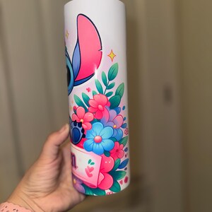 May include: White tumbler featuring a colorful illustration of a blue alien character with large ears, surrounded by pink, blue, and purple flowers. The design includes a banner with text. The tumbler is held by a person.