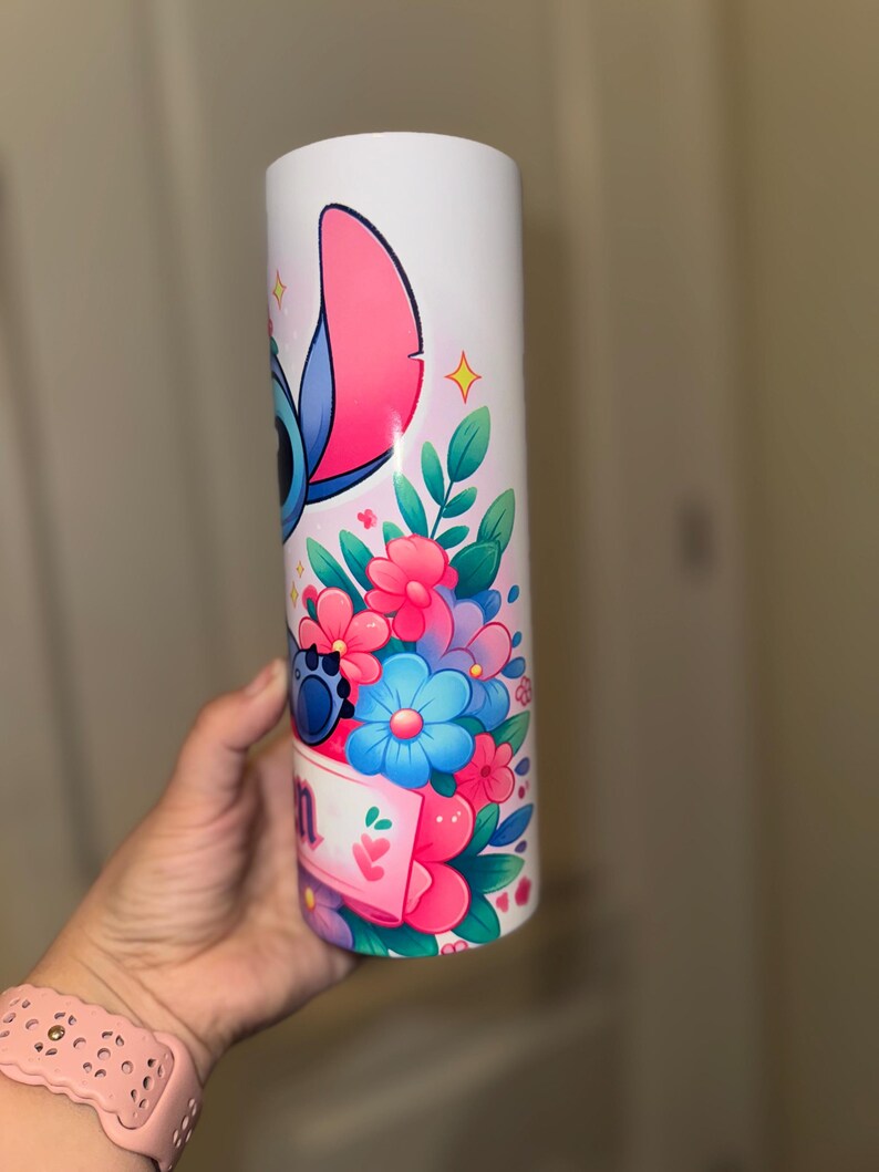 May include: White tumbler featuring a colorful illustration of a blue alien character with large ears, surrounded by pink, blue, and purple flowers. The design includes a banner with text. The tumbler is held by a person.