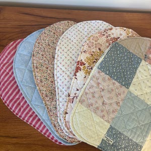 May include: A set of six oval quilted fabric placemats featuring various patterns including red and white stripes, solid light blue, small floral prints, and a patchwork design. A small white label reads Clover & Craft Studio.