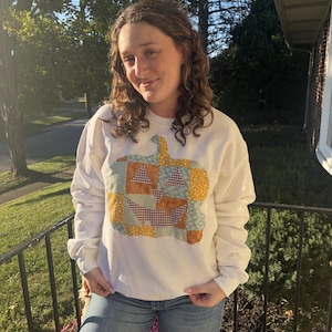 May include: A white long-sleeve sweatshirt featuring a large, central pumpkin graphic constructed from a patchwork of various fabric patterns in shades of orange, yellow, blue, and brown, paired with blue denim jeans.