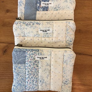 May include: Three rectangular zippered pouches made of quilted fabric in various shades of blue and white floral and geometric patterns. Each pouch features a small white fabric label with the text Clover & Craft Studio stitched on the front.