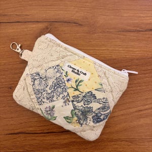 May include: A quilted zipper pouch featuring a patchwork design of cream, yellow, and blue floral fabrics. The pouch includes a white zipper, a silver metal lobster clasp on a side tab, and a white fabric label reading Clover & Craft Studio.