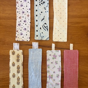 May include: A set of seven rectangular fabric bookmarks with lace tabs. Patterns include purple pumpkins and stars, light blue paisley, multicolored dots, yellow floral prints, solid light blue, purple roses, and a red and white gingham check.