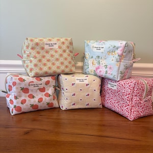 May include: Five fabric cosmetic bags with various floral and fruit patterns. The bags feature a strawberry, rose, and cherry design. Each bag has a zipper closure and a small tag that reads "Clover & Craft Studio". The bags are on a wooden surface.