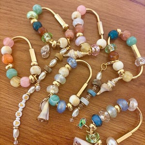 May include: A collection of gold-toned metal keychains featuring assorted colorful beads in shades of blue, pink, orange, and green. Each keychain includes small seashell charms and one keychain features a dangling strand with white letter beads spelling MAMA.