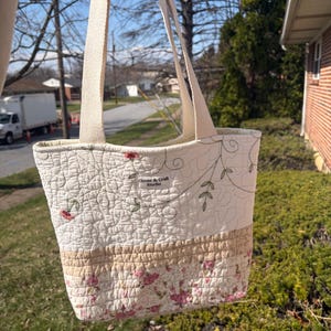 May include: Cream-colored quilted tote bag with floral and vine patterns. The bag features a beige bottom section and cream-colored handles. The text "Clover & Craft Studio" is visible on the side of the bag. The bag is held up in front of a sunny outdoor scene.