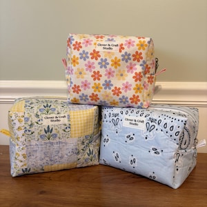 May include: Three rectangular zippered pouches with quilted fabric. The top pouch features a white background with orange, pink, and blue flowers. The bottom left pouch has a blue and yellow floral patchwork pattern. The bottom right pouch is light blue with a white paisley print. Each pouch has a white label reading Clover & Craft Studio.