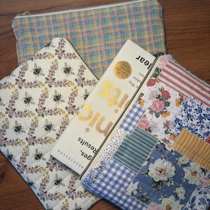 May include: Three zippered pouches in various patterns. One is plaid, another features bees and floral designs, and the third is a patchwork of floral and gingham fabrics. A book with gold lettering is partially visible.