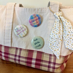 May include: A beige canvas tote bag with a red and beige plaid bottom panel. The bag features three circular pin-back buttons with text: "I LOVE A TRINKET AND A TREAT", "BORN TO DILLY DALLY", and "hot girls craft". A floral fabric ribbon is tied to the handle.