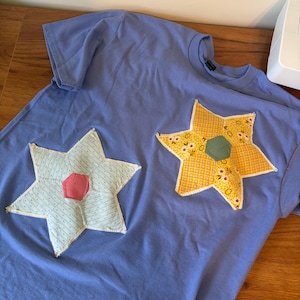 May include: A light blue t-shirt with two star-shaped appliqués. One star is light blue with a pink center, and the other is yellow with a green center. The shirt is lying on a wooden surface.
