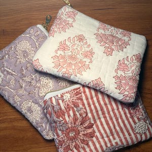 May include: Three quilted pouches with floral and striped patterns. One pouch is lavender with white floral designs, another is white with pink floral designs, and the third is red and white striped with floral accents. Each has a gold zipper.