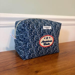 May include: A quilted fabric pouch in dark blue with a white paisley pattern. It features a gold zipper, a white label reading Clover & Craft Studio, and an oval patch with a red border, three blue stars, and the text American Made in blue.