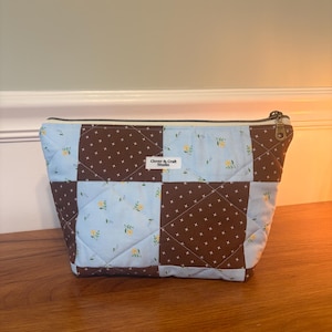 May include: A quilted zippered pouch featuring a patchwork design of light blue fabric with yellow floral patterns and dark brown fabric with a white cross-stitch pattern. A white label with the text Clover & Craft Studio is centered on the front.