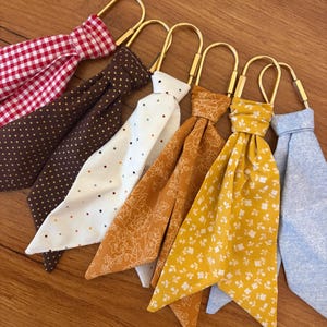 May include: Six fabric keychains featuring gold-toned metal U-shaped rings. The fabric patterns include red and white gingham, brown with gold dots, white with multicolored dots, mustard yellow with floral line art, mustard yellow with white flowers, and light blue.