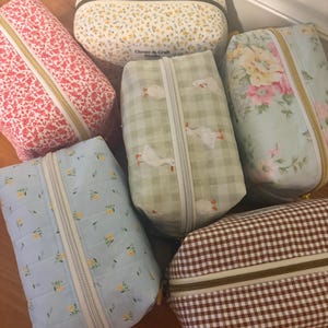 May include: A collection of six rectangular fabric pouches with zippered closures. The pouches feature various patterns including red floral, yellow floral, green gingham with white geese, blue floral, brown gingham, and a light blue floral print with quilting.