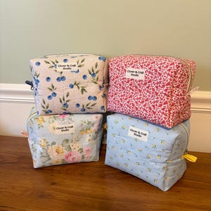 May include: Four rectangular quilted fabric pouches with zippered closures. Each pouch features a white label reading Clover & Craft Studio. Patterns include blue berries, red floral, pink and yellow roses, and small yellow flowers on light blue.