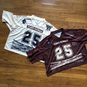 May include: Two American football jerseys, one cream and one maroon, are shown on a wooden surface. Both jerseys have "CHRIS BROWN" above the number "25" in white. The cream jersey has black numbers, the maroon jersey has white numbers.