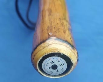 May include: Close-up of a wooden walking stick with a compass embedded in the handle. The compass face is white with black markings and a red needle pointing north. The stick is brown with a natural wood finish.