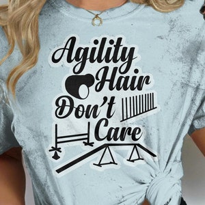 May include: A light blue t-shirt with a distressed look featuring the text "Agility Hair Don't Care" in black. The text is surrounded by a black illustration of an agility course.