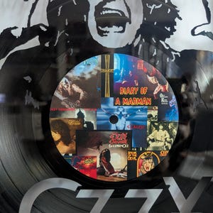 May include: Black vinyl record with a collage of album covers in the center, featuring the text "Diary of a Madman." The record is set against a black and white graphic of a face and the word "OZZY" in silver.