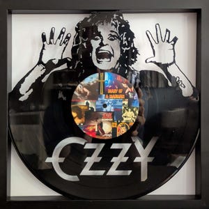 May include: Framed black vinyl record featuring a silhouette of a screaming figure with outstretched hands. The record has the name "OZZY" in white lettering and a central image of album covers. The frame is black.