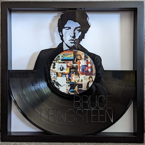May include: A black vinyl record with a silhouette of Bruce Springsteen's head and a collage of album covers on the record. The record is framed in a black frame with a white mat.