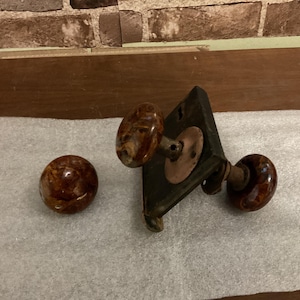 May include: Two brown ceramic door knobs with a metal door plate and a metal connecting rod.