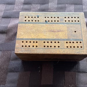 May include: A vintage wooden cribbage board with multiple rows of drilled holes for pegging. The board is rectangular with a natural wood finish, accented by thin, dark blue lines. The surface shows signs of wear, suggesting age and use.
